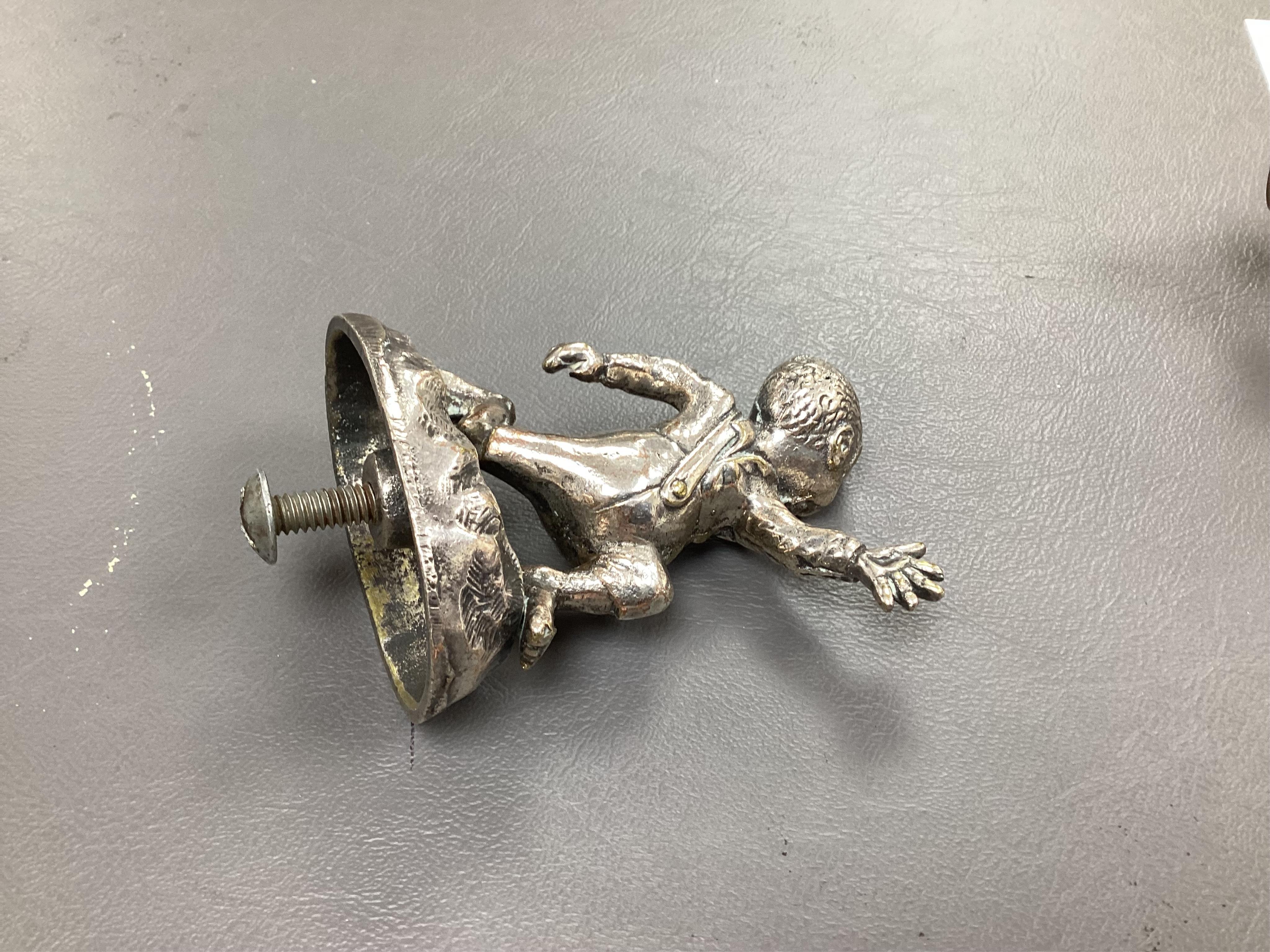 Seven chrome and brass car mascots, including a Mercedes-Benz logo, a figure of a blacksmith, a caricature figure on a beer barrel, a golf ball in mid flight, etc. together with a chromed housing unit for an electric lam